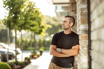 handsome man with short bristles in a black T-shirt leans against a brick wall