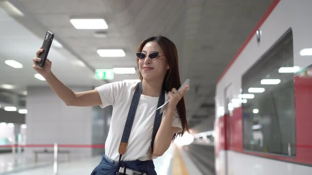 Selective focus of a happy beautiful Asian female traveler with a film camera hanging on neck, wearing sunglasses, using a cellphone taking selfie with a blurred stationary train at a subway platform.