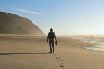 man walking along a deserted beach with a camera in his hand, back view.
