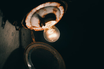 An old incandescent light bulb glows in the basement in an old glass shade with a rusty base