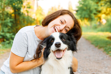 Smiling young attractive woman playing with cute puppy dog border collie on summer outdoor background. Girl holding embracing hugging dog friend. Pet care and animals concept - obrazy, fototapety, plakaty