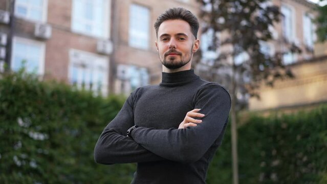 Portrait of ambitious confident Caucasian young man crossing hands looking at camera. Smiling brunette guy posing outdoors standing on city street. Confidence and lifestyle concept