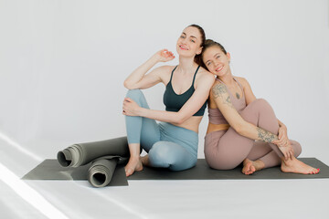 The girls smile and rest after yoga classes on the mat leaning on each other. Sunbeams illuminate the white studio