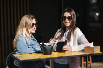 Two beautiful blonde and brunette girls order takeaway chinese food. Young woman outdoors speaking. Spring sunny day