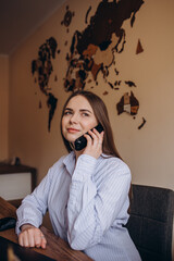 Concept for travel agent. Young blonde woman smiling, looking at camera and using mobile phone. Travel agency office interior with big world map and window