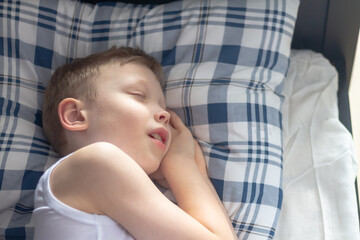A blond Caucasian boy sleeps on a bed on blue checkered pillows and under a blanket