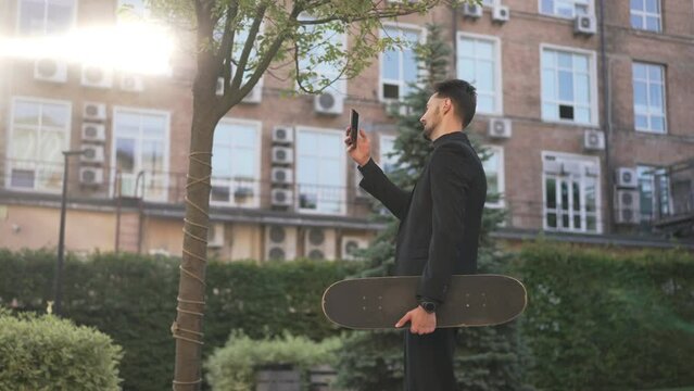 Side view young smiling man with skateboard walking in city at sunrise surfing social media on smartphone. Live camera follows happy Caucasian guy in suit strolling in the morning in slow motion