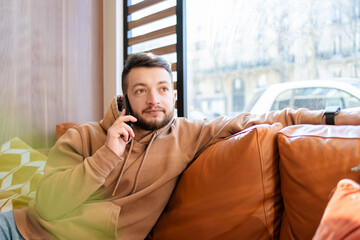 Smiling man using smartphone and phoning on sofa at window. Handsome young guy working at cafe,...