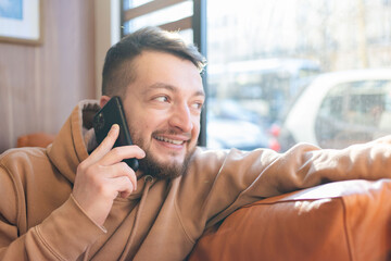Portrait of an attractive young bearded man wearing casual clothes sitting on a couch at the cafe, talking on mobile phone, chatting, messaging.