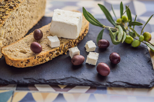 Green and black olives with loaf of fresh bread, feta cheese and young olives branch on olive wood chopping board over dark background - Powered by Adobe