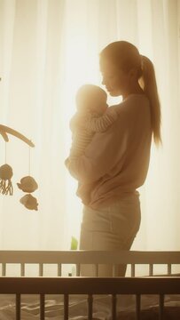 Vertical Format Portrait Video of Happy Mother Holding Cute Newborn Baby in Cozy Dark Kids Bedroom in the Evening. Caring Mom Hugging the Neonate Toddler and They are Bonding Before Sleep.