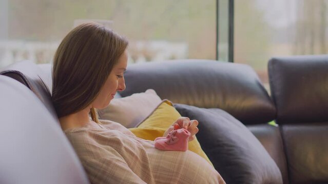 Pregnant woman sitting on sofa at home looking a small baby shoes  - shot in slow motion