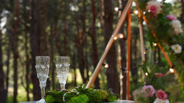 Wine glasses, wedding rings bedding of grass and moss on white table next to bohemian tipi arch decorated in boho style with flowers and candles and white chairs for guests.