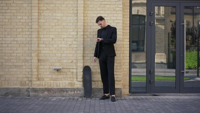 Wide shot portrait of confident Caucasian businessman standing on city street with skateboard checking time on wristwatch. Serious elegant handsome man waiting for meeting in urban town. Slow motion