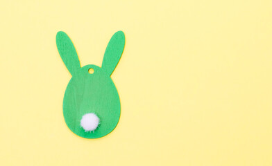 A green wooden decorative Easter bunny with a white fluffy round ponytail on a yellow background.