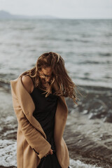 young woman of Slavic appearance walks on the beach of the sea, dressed in a beige coat and black dress, emotions on her face, smile, euphoria, joy, blond hair