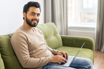 Young Indian male student watching webinars, educational courses, learning on the distance. Front view of positive Hindi man in smart casual clothes using laptop while sitting at the sofa in his flat