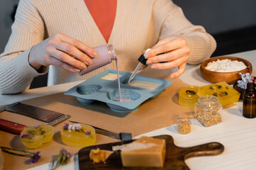 Fototapeta na wymiar Cropped view of craftswoman pouring soap in mold near flowers, cutter and flakes on table.