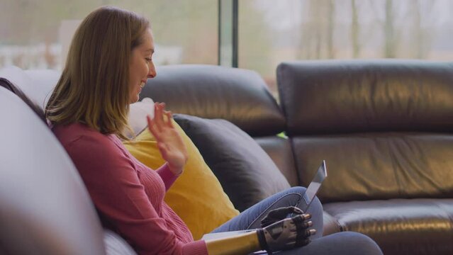 Side view of young woman with prosthetic arm and hand sitting on sofa at home waving as she makes video call using laptop - shot in slow motion