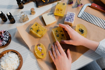 Top view of craftswoman holding handmade soap on craft paper near cutter and flowers on table.