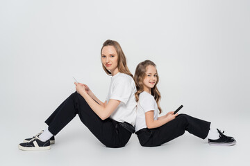 woman and child with mobile phones sitting back to back and looking at camera on grey background.