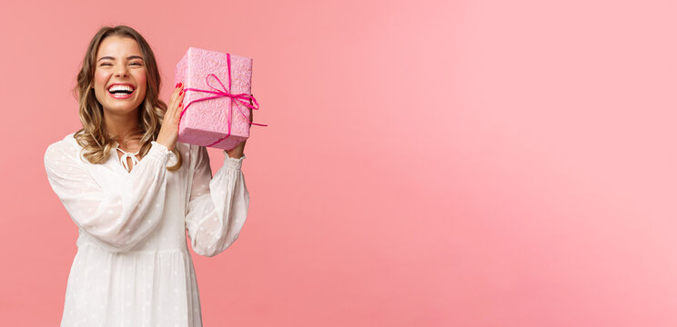 Holidays, Celebration And Women Concept. Portrait Of Happy Charismatic Blond Girl Shaking Gift Box Wondering Whats Inside As Celebrating Birthday, Receive B-day Presents, Pink Background