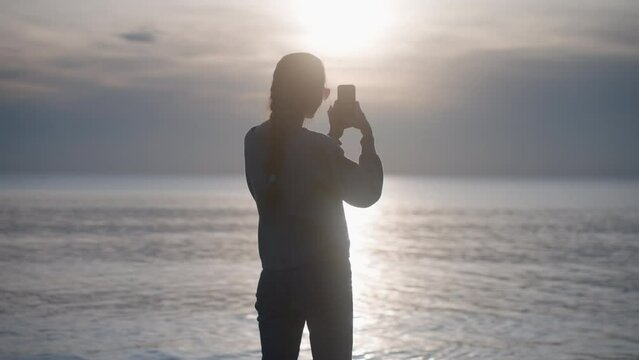 Silhouette of young woman shooting sea and amaizing sunset view on smartphone. Beautiful stylish brunette with braid and sunglasses takes photo of seaside landscape