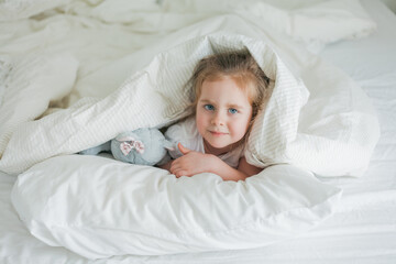 Little cute blue-eyed curly girl plays in bed with her plush toy. The child is lying in bed under the covers. Emotions. Healthy sleep.