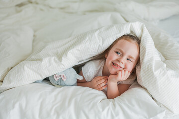 Little cute blue-eyed curly girl plays in bed with her plush toy. The child is lying in bed under...
