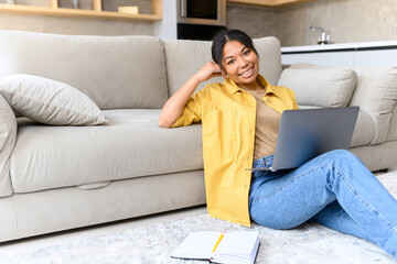 Cheerful serene African-American young woman using laptop, smiling girl sitting on the floor and looking at the camera, attractive female student studying on the distance