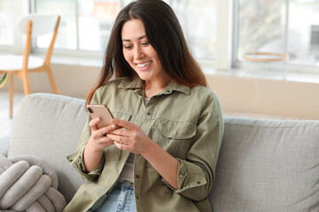 Beautiful young Asian woman with mobile phone sitting on sofa at home
