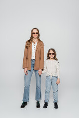 full length view of trendy mother and daughter in sunglasses holding hands on grey background.