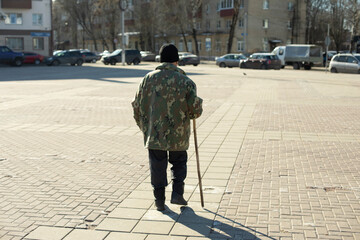 Grandpa walks through square. Elderly man in town. Man with stick. Pensioner on street.