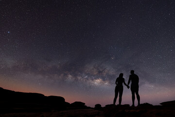 Milky Way with silhouette of people. Landscape with night starry sky. Standing man and woman on the mountain with yellow light. Beautiful galaxy. Universe.