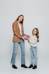 full length view of trendy mother and daughter smiling near gift box on grey background.