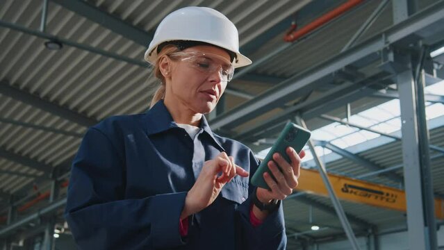 Close up young woman with helmet uses a mobile phone in factory. In the background details of a metalworking project