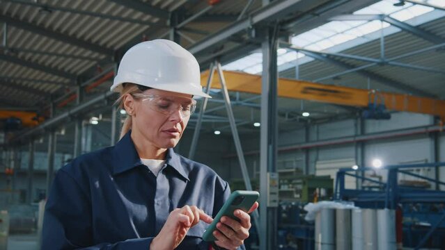 Serious woman engineer with helmet and goggles uses mobile phone in factory. In the background details of a metalworking project