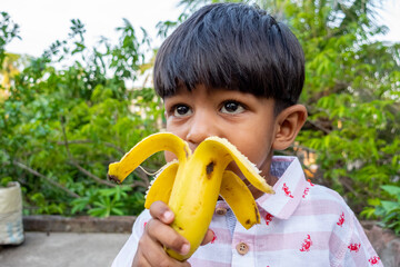 A boy is standing in the garden and eating a ripe banana. Boy eating food. Asian boy eating banana.