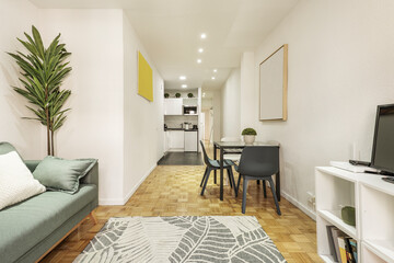 Living room of a vacation rental apartment with a tv on a white shelf, a sofa upholstered in light green fabric, a metal and square glass dining table and wooden floors