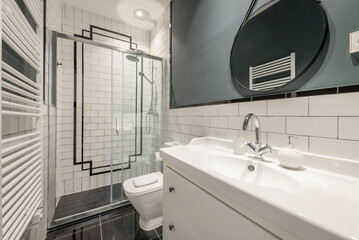Bathroom with white porcelain sink and contemporary decoration with round mirror with black frame, shower cabin with glass screen and white tiles