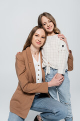 joyful woman and stylish girl smiling at camera while embracing isolated on grey.