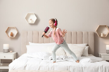 Cute little girl in headphones with hairbrush singing on bed at home