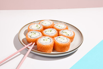 Fototapeta na wymiar Philadelphia maki roll on ceramic plate with chopsticks. Salmon sushi roll with philadelphia cheese on coloured background. Japanese menu concept. Salmon sushi roll in modern style.