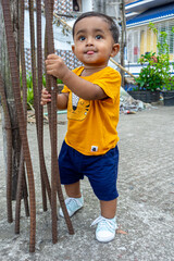 A infant child is trying to stand up holding a rusty iron rod in his small two hands. A kid is having fun and sticking out his tongue.