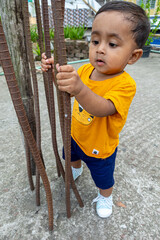 A infant child is trying to stand up holding a rusty iron rod in his small two hands. A kid is having fun and sticking out his tongue.