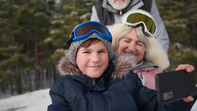 pensioners with grandson in forest, smiling male child filming video on smartphone camera together with grandparents and sledding from snowy hill among outdoor trees