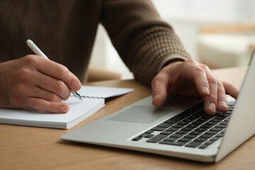 Fototapeta premium Man with laptop and notebook learning at wooden table indoors, closeup