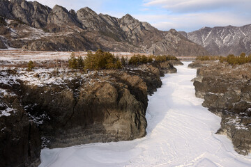 Frozen riverbed squeezed into rocky high banks flowing through a snow-covered valley surrounded by mountain ranges in winter.