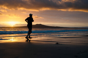 Fototapeta premium Silhouette man running on Takapuna beach at sunrise, Rangitoto Island in the background, Auckland.