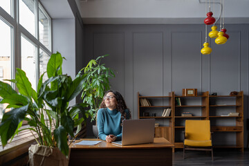 Satisfied happy female employee relaxing at workplace in modern office, sitting in chair in front of laptop, receiving good news. Businesswoman enjoying break during workday. Happiness at work concept